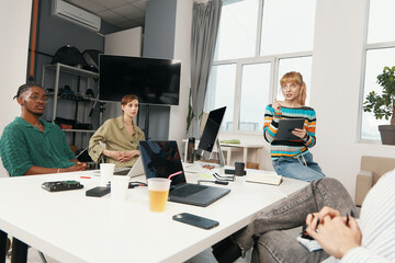 Young adult woman leading business meeting with diverse colleagues working and socializing in...
