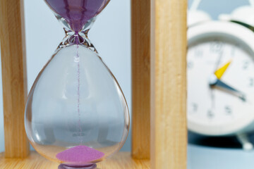 Hourglass with purple sand flowing, showing the passage of time, with a blurred alarm clock in the...