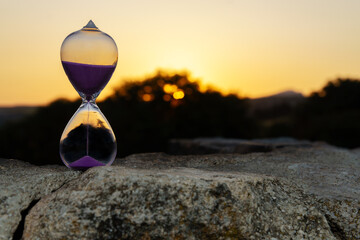 Hourglass sits on a rock, measuring the passing time as the sun sets behind a blurred natural...