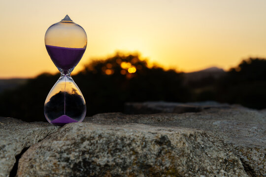 Hourglass sits on a rock, measuring the passing time as the sun sets behind a blurred natural landscape