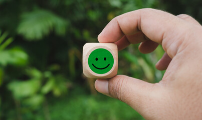 Mental well-being with nature, tranquility in the forest, mental health care, World Mental Health Day, Fresh Green Leaves, Hand holding wood cube with smile icon on green forest, Positive vibe hiking