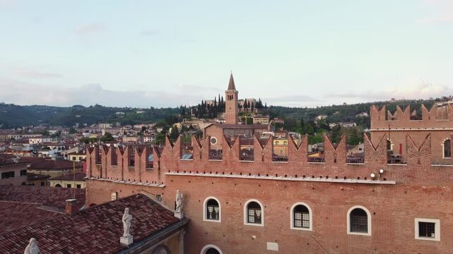 Drone aerial of Verona shows the brick Gothic Palazzo del Podest&agrave; with its battlements, and the tall bell tower of San Zeno in the distance, surrounded by hillside cypresses, slow glide, wide shot