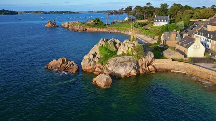 Drone arrives from the sea toward the Sentinelle oratory, showing houses, bay, rocks and calm coastal waters. Port Blanc, Bretagne - France