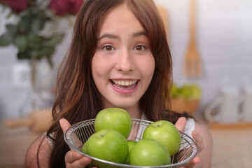 Close up, happy teenage girl holding basket full of green apples in home kitchen, looking positive...