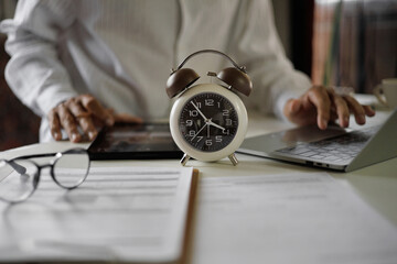 selective focus on alarm clock, asian businessman using laptop and tablet working on table before work time end in a day,  time management concept, routine work, office hour, blur background