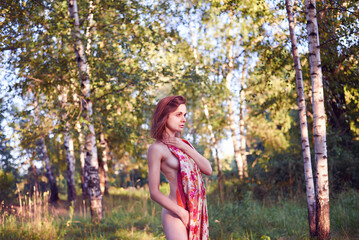 A woman is standing in a field with a red scarf around her neck. She is bare-breasted and she is...