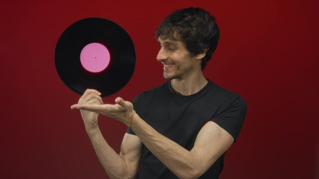 Young man smiling holds vinyl record by pink label on fingers in red studio; nostalgia music art vintage.