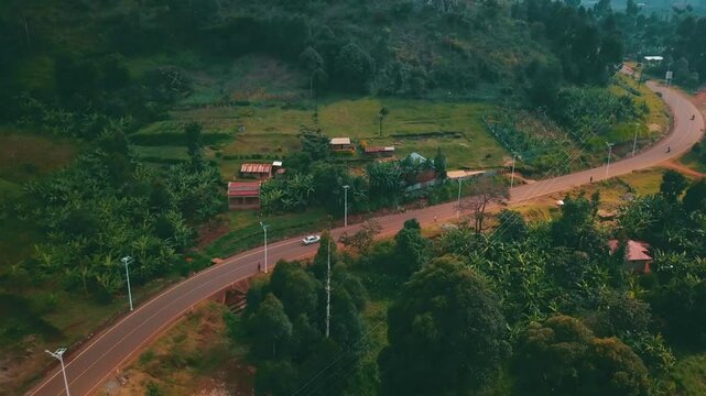 Drone aerial of a white car traveling a curved road surrounded by trees, banana crops, and rural buildings on Mount Elgon&rsquo;s fertile slopes in Eastern Uganda