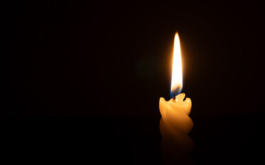 A single burning candle flame or light glowing on a spiral white candle on black or dark background on table in church for Christmas, funeral or memorial service with copy space