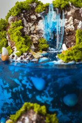 Beautiful birthday cake in the shape of an island decorated with chocolate edible mountains, greenery with waterfall. Tropical island cake surrounded with blue jelly ocean water