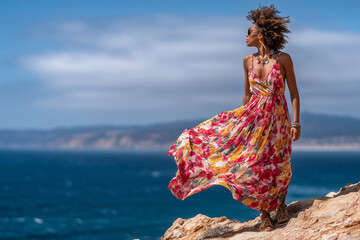 Woman in flowy floral dress enjoying wind on seaside cliff, 