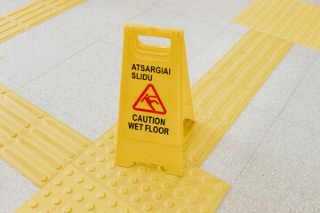 Yellow plastic caution sign on tactile paving for visually impaired, warning about wet floor in a...