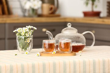 Wooden tray with teapot, cups of natural chamomile tea and flowers on table in kitchen
