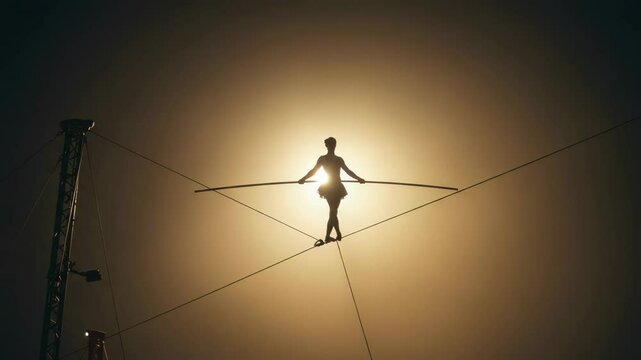 Tightrope walker balancing against illuminated backdrop at sunset  