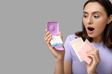 Beautiful shocked woman with tampons storage box and menstrual pads on grey background