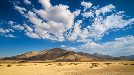 A vast, sandy desert landscape stretches to meet a dramatic mountain range under a brilliant blue sky scattered with fluffy white clouds