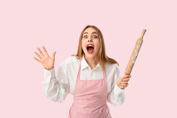 Surprised young woman in apron with rolling pin on pink background