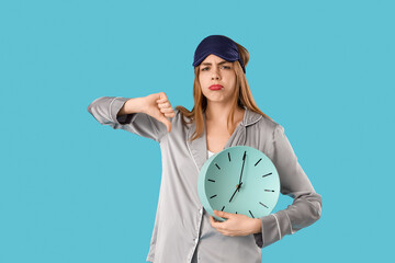 Portrait of displeased young woman in pajamas with clock showing thumb-down on blue background