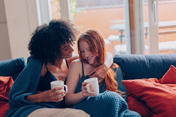 Two smiling women are hugging and drinking coffee on the sofa