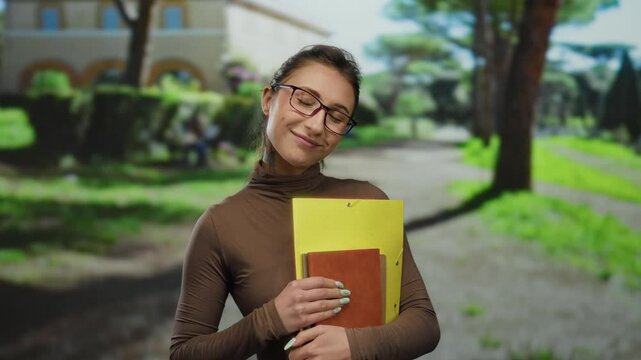 Young woman with glasses holding folders smiling in a sunlit park setting showcasing joy creativity and learning outdoors surrounded by lush greenery and a clear blue sky.