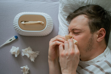 Sick man blowing his nose in bed, suffering from cold or flu, with a thermometer and used tissues...