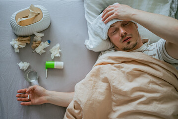 Sick man lying in bed with a wet towel on his forehead, surrounded by tissues, medication, and a...