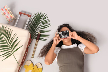 Young African-American woman lying with travel accessories and using camera on grey background. Top view