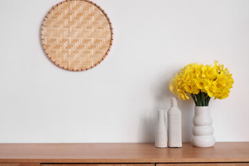 Vase with narcissus flowers on chest of drawers near white wall in light room