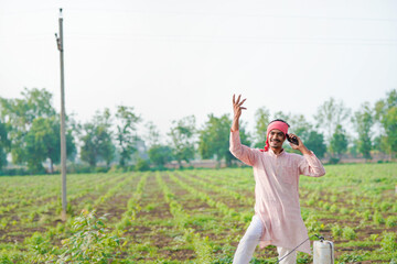 young indian farmer standing at field talking on smartphone