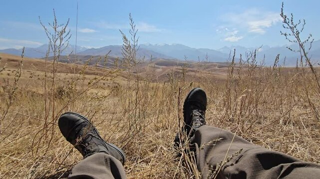 First-person perspective POV shot of legs in military combat boots resting in dry grass, enjoying the mountain landscape view in Kyrgyzstan.
