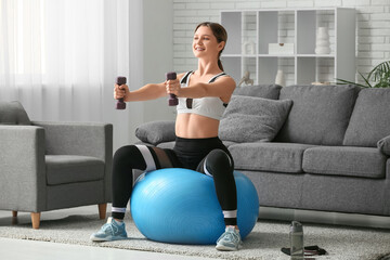 Beautiful young woman training with dumbbells on fitball in living room