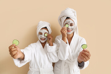 Happy young African-American mother and her daughter in bathrobes with facial clay masks applying cucumber slices on beige background