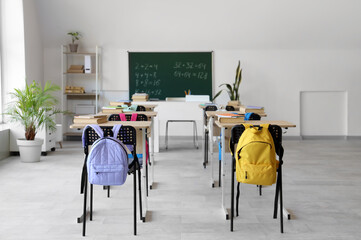 Interior of light classroom with desks, backpacks on chairs and blackboard