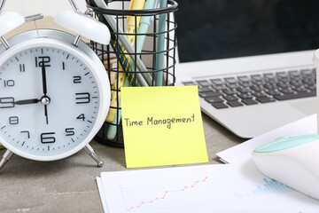 Sticky note with text TIME MANAGEMENT, alarm clock,gadgets and stationery on table, closeup