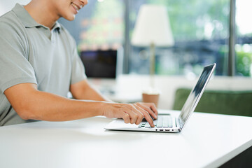Young man typing on laptop and smartphone at modern office desk with coffee and tablet nearby.