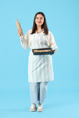 Young woman holding baking tray with cookies and rolling pin on blue background