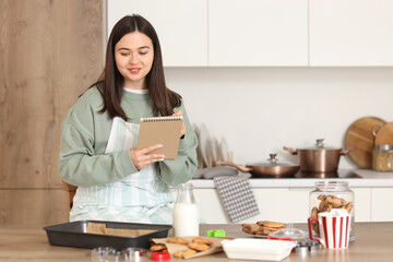 Young woman with cookies writing on notebook at table in kitchen