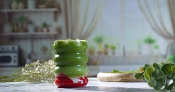 A hand gently places a vibrant layered matcha strawberry latte on a rustic white table, surrounded by greenery and soft natural light. Ideal for beverage ads and social media content.
