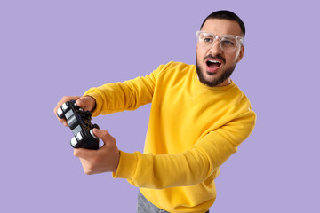 Emotional young man in eyeglasses playing video games on lilac background