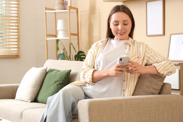 Beautiful young woman using modern mobile phone on stylish grey sofa in living room