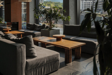 Sunlit hotel lounge with grey modular sofas, solid wood coffee tables and potted greenery, framed by panoramic windows overlooking mountain-side resort buildings