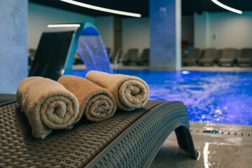 Rolled beige towels on a wicker lounger beside an illuminated indoor pool with a cascading water...