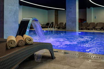 Rolled beige towels on a wicker lounger beside an illuminated indoor pool with a cascading water...