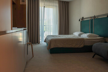 Minimalist hotel bedroom with teal upholstered headboard, double bed neatly made, floor-to-ceiling...