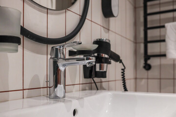 Close-up modern hotel bathroom sink with shiny chrome faucet, oval mirror and wall-mounted...
