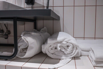 Rolled white terry bathrobes and fresh towels neatly stored beneath modern hotel bathroom sink,...
