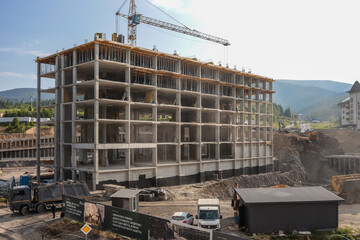 Concrete skeleton of multi-storey building rises beside a tower crane on busy mountain-resort construction site; dump truck and excavators dig foundations against a clear summer sky