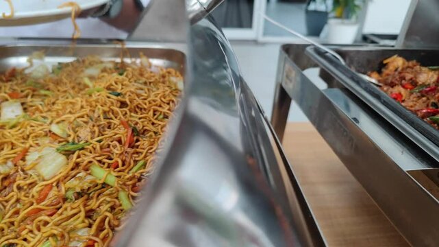 Cinematic footage of a buffet spread featuring classic Asian dishes, including stir-fried noodles (mie goreng) and spicy seasoned chicken (ayam bumbu pedas). The food is presented in stainless steel 