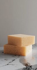 Two light beige sponges stacked on a marble surface against a light gray background