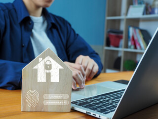 Wooden house model with security lock icon and fingerprint symbol near person typing on laptop...